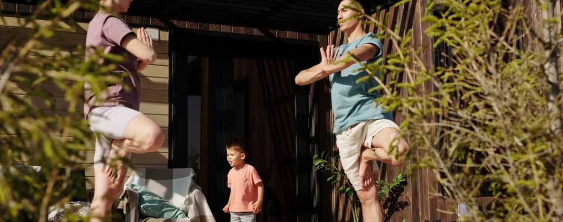A family practicing yoga in tree pose on a sunlit porch, promoting health and mindfulness.